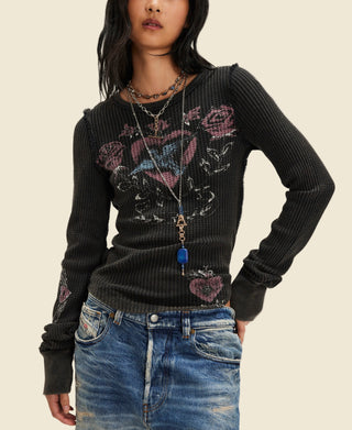 A model wearing a black ribbed knit long-sleeve sweater with a distressed graphic of a heart, roses, and a dagger on the front, paired with blue jeans and layered necklaces, against a plain white background.