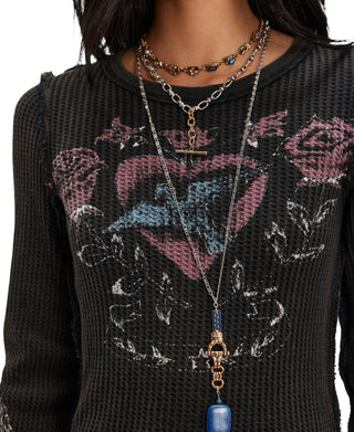 A close-up of a person wearing layered necklaces over a black mesh long-sleeve shirt with a pink, blue, and white graphic print. The necklaces include a silver chain with a gold-tone T-bar clasp and a blue bead pendant, a gold-tone chain with a skull charm, and a beaded choker.