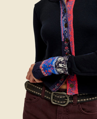 A woman wearing a black long-sleeve knit noa cardigan from free people with a colorful vertical patterned placket and matching patterned cuff trim, paired with dark brown pants and a studded belt, against a plain white background.
