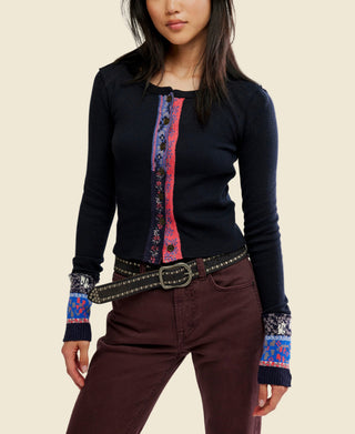 A woman wearing a black long-sleeve knit noa cardigan from free people with a colorful vertical patterned placket and matching patterned cuff trim, paired with dark brown pants and a studded belt, against a plain white background.